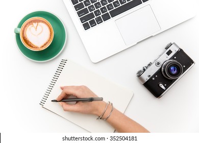 Top View Of Laptop, Old Camera, Cup Of Coffee And Woman Hand Writing In Notebook Over White Background