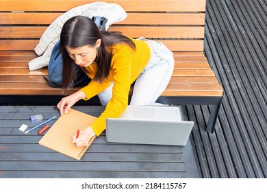 Top View Of A Latin Student Woman Wearing A Yellow Sweater Taking Notes In Her Notebook Next To Her Laptop On A Wooden Bench Outside