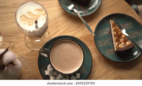 Top view of latte with intricate foam art, accompanied by a cup of hot chocolate on a green saucer and a slice of caramel nut cheesecake, on a wooden table setting. - Powered by Shutterstock