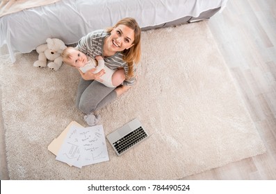 Top View Of Laughing Mother Sitting On Tapis And Holding Calm Baby. They Are Looking At Camera. Copy Space In Right Side