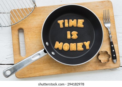 Top View Of Letter Collage Made Of Biscuits. Quote TIME IS MONEY In Black Frying Pan. Other Cooking Equipments: Fork, Cookie Cutter And Chopping Board Putting On White Wood Table, Vintage Style Image.