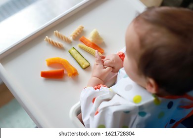 Top View Of Little Boy Holding Food In His Hands Baby Led Weaning (BLW) With Mixed Vegetables And Pasta On High Chair. Happy Infant Eating Self Feeding.