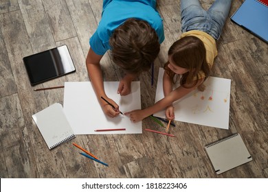 Top View Of Little Caucasian Boy And Girl Lying On The Wooden Floor At Home And Drawing On A White Sheet Of Paper With Colorful Pencils