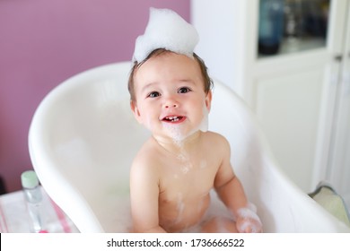 Top view of little cute baby take a bath and playing with foam bubbles with smiling face.mixed race Asian-German infant bathing in white bathtub laughing and fun. Happy child in bathroom.