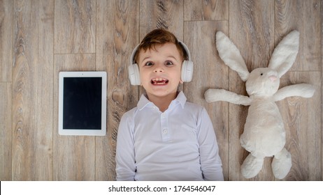 Top View Of Little Kid Boy Laying On The Floor With His Rabbit Toy And Listen To The Music In Wireless Headphones.