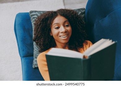 Top view little smart kid teen girl wear casual clothes reading novel paper book sits on blue sofa couch stay at home hotel flat rest relax spend free spare time in living room indoor. Lounge concept - Powered by Shutterstock