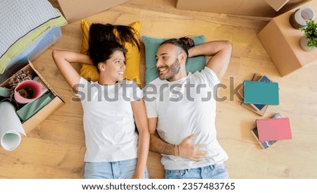 Top view of loving cheerful millennial couple have break while unpacking stuff after moving to new house, lying on floor among packed belongings, looking at each other and smiling