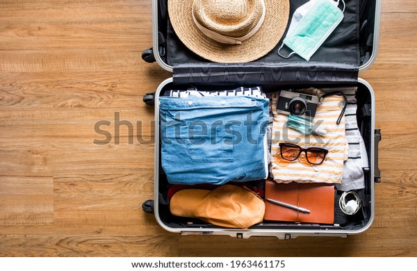  Top view of Luggage,suitcase bag with clothing and\
mask on wood floor.summer and vacation with coronavirus\
pandemic.copy space