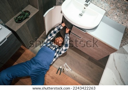 Top view. Lying down on the ground, fixing water tap. Plumber in blue uniform is at work in the bathroom.