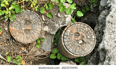 Top view macro of rusty cast iron PDAM water meter lids surrounded by soil and green wild plants