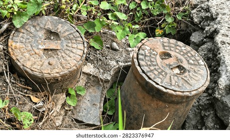 Top view macro of rusty cast iron PDAM water meter lids surrounded by soil and green wild plants