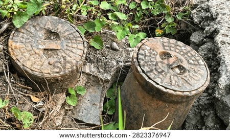 Top view macro of rusty cast iron PDAM water meter lids surrounded by soil and green wild plants