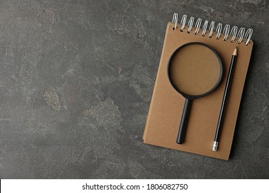 Top view of magnifier glass, empty notebook and pencil on grey stone background, space for text. Find keywords concept