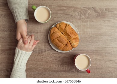 Top View Of Male And Female Holding Hands Over The Table With Two Cups Of Coffee And Plate Of Croissants Is Placed Next To It