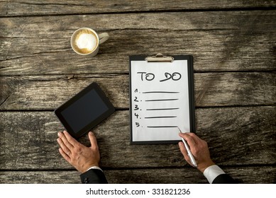 Top View Of Male Hand Writing A To Do List On A White Sheet Of Paper With Digital Tablet And Cup Of Coffee Alongside On His Textured Rustic Wooden Desk.