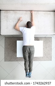 Top View Of Male Person Laying On Sofa And Coffee Table. Man Trying To Push Back Grey Couch. Fluffy Carpet On Floor. Light Interior And Renovation Concept