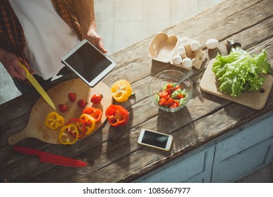 Top View Of Male Person Standing At The Kitchen Table With Tablet And Knife In Hands. Vegetables And Gadget Are On Desk