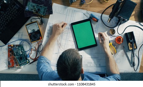 Top View Of Male IT Technician Working On Blueprints, He's Tapping On A Green Screen Tablet Computer. He's Surround By Circuit Boards, Laptop And Other Technical Components. Sun Shines On His Table.