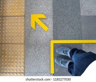 Top View Of Man Feet Standing Over Arrow Symbol On Subway Platform. Yellow Arrow Sign On Floor At The Train Station.