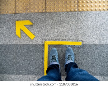 Top View Of Man Feet Standing Over Arrow Symbol On Subway Platform. Yellow Arrow Sign On Floor At The Train Station.