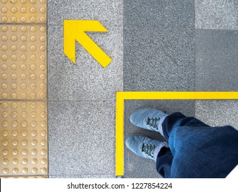 Top View Of Man Feet Standing Over Arrow Symbol On Subway Platform. Yellow Arrow Sign On Floor At The Train Station.