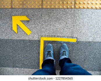 Top View Of Man Feet Standing Over Arrow Symbol On Subway Platform. Yellow Arrow Sign On Floor At The Train Station.