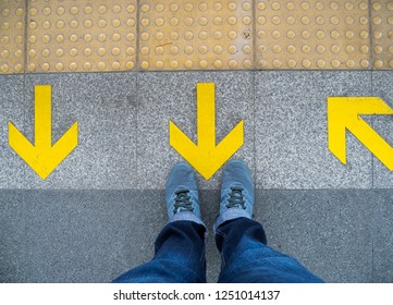 Top View Of Man Feet Standing Over Arrow Symbol On Subway Platform. Yellow Arrow Sign On Floor At The Train Station.