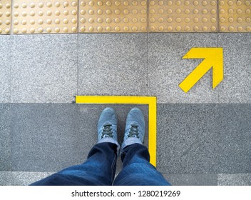 Top View Of Man Feet Standing Over Arrow Symbol On Subway Platform. Yellow Arrow Sign On Floor At The Train Station.