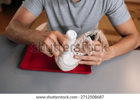 Top view of man using bottle of shoe whitener and painting scuffed surface of dirty white sneaker, illustrating process of cleaning, recoloring, and restoring footwear.