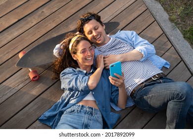 Top view of man and woman with skateboard watching video or photos on smartphone. Smiling boyfriend and girlfriend lying on wooden surface and enjoying time together. Relationship, entertainment idea - Powered by Shutterstock