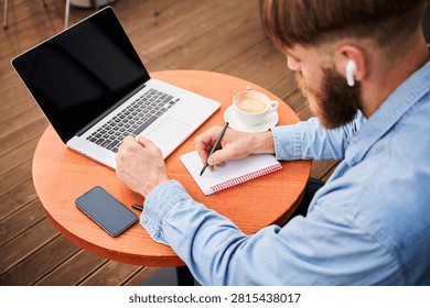 Top view of man writing in notebook beside laptop, coffee cup, and smartphone at outdoor terrace table, showing focused remote work and planning routine