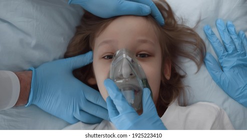 Top View Of Medical Team In Protective Gloves Putting Oxygen Mask On Patient Child In Hospital Bed. Close Up Of Doctors Reanimating Sick Kid In Hospital Ward