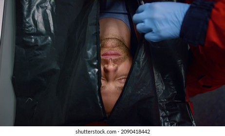 Top View Medical Worker Hands Closing Black Bag With Dead Man Inside. Closeup Paramedic In Gloves Zipping Up Body Bag With Corpse. Cadaver In Plastic Bag Lying On Stretchers In Ambulance Car