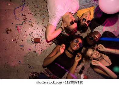 Top View Of Men And Women Lying On Floor With Heads In The Centre And Laughing At A House Party. Friends Lying On Floor At The House Party With Cups And Confetti Around.