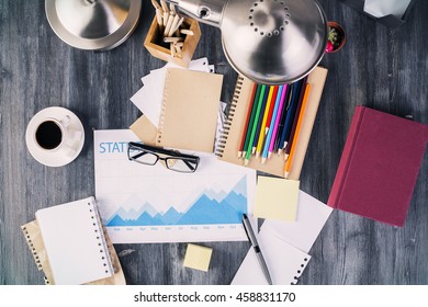 Top View Of Messy Desktop With Business Report, Coffee Cup, Table Lamp, Book And Various Stationery Items