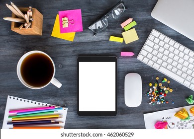 Top View Of Messy Wooden Desktop With Blank White Tablet, Computer Mouse And Keyboard, Mug With Tea, Glasses, Colorful Stationery And Office Tools. Mock Up