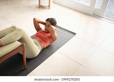 Top view of mid adult fit woman doing press on a mat while keeping legs on chair at home. African american woman practicing crunches at home. High angle view of healthy mixed race girl doing abs. - Shutterstock ID 1999270205