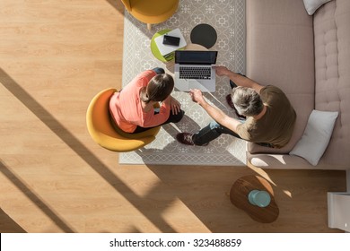 Top View Of A Middle Age Man And Woman In Casual Clothes. They Are Sitting In A Stylish Vintage Living Room With Wooden Floor The Grey Hair Man Is Showing Some New Project On The Computer To The Woman