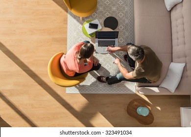 Top View Of A Middle Age Man And Woman In Casual Clothes. They Are Sitting In A Stylish Vintage Living Room With Wooden Floor The Grey Hair Man Is Showing Some New Project On The Computer To The Woman