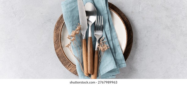 Top view of a minimal and elegant table setting featuring a ceramic plate with blue linen napkin, wooden-handled cutlery, and glassware on a light textured background. Dried flowers add a rustic touch. Perfect for food, lifestyle, or hospitality themes.