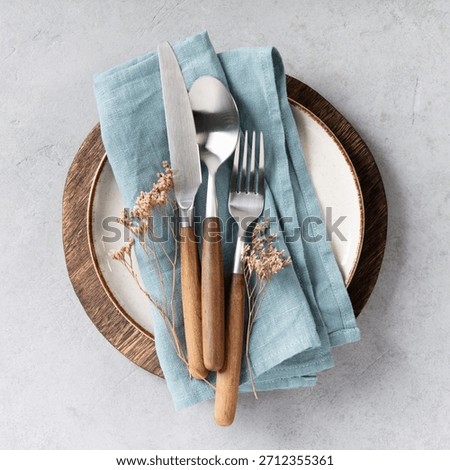 Top view of a minimal and elegant table setting featuring a ceramic plate with blue linen napkin, wooden-handled cutlery, and glassware on a light textured background. Dried flowers add a rustic touch. Perfect for food, lifestyle, or hospitality themes.