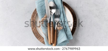Top view of a minimal and elegant table setting featuring a ceramic plate with blue linen napkin, wooden-handled cutlery, and glassware on a light textured background. Dried flowers add a rustic touch. Perfect for food, lifestyle, or hospitality themes.