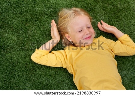Top view. Mock up for logo, text, design. Blonde long hair child girl lying stretching on green grass. Preschool girl 5-6 years old in yellow t shirt. Lifestyle Summer vacation Leisure. People