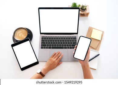 Top view mockup image of hands holding a blank white screen mobile phone with laptop computer and tablet pc on the table in office