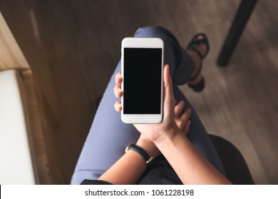 Top view mockup image of a woman holding white mobile phone with blank black desktop screen on thigh in cafe