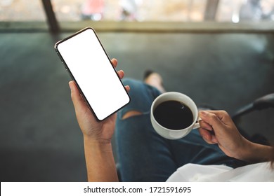 Top View Mockup Image Of A Woman Holding A Black Mobile Phone With Blank White Desktop Screen With Coffee Cup