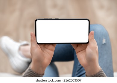 Top View Mockup Image Of Woman Holding Mobile Phone Horizontally With Blank White Screen. Woman In Jeans Sits Cross-legged And Holds Cell Phone Horizontal In Hands With Blank White Screen