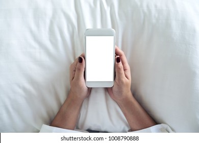 Top View Mockup Image Of A Woman's Hand Holding White Mobile Phone With Blank Desktop Screen On Pillow And Sitting In The Bed