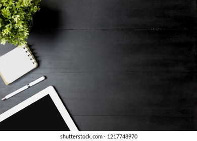 Top View Modern Black Office Wood Desk Table. Workspace With Blank Tablet, Pen, Notebook, Plant Mockup And Black Background