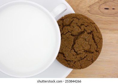 Top View Of A Molasses Cookie On A Saucer With Whole Milk In A Coffee Cup Atop A Wood Table Top.